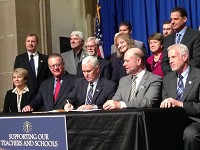 Gov. Mike Pence signs legislation Thursday that holds harmless schools and teachers from negative effects tied to poor student performance on the 2015 ISTEP exam. Glenda Ritz, the state superintendent of public instruction, stands directly behind the governor as numerous state lawmakers and Lt. Gov. Sue Ellspermann, seated at far left, look on. Staff photo by Dan Carden