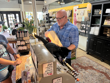 Richard Sanders, owner of Ditto’s on Main in Madison, checks a customer out while shop cat Dash seeks affection. (Niki Kelly/Indiana Capital Chronicle)